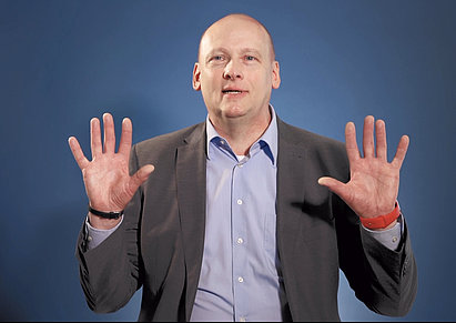 Man in a suit with shaved head raising both hands with palms facing forward against a blue background